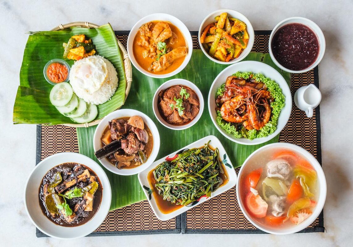 This image showcases a vibrant spread of Peranakan or Southeast Asian dishes, featuring a variety of curries, stir-fried vegetables, and sambal prawns served alongside rice topped with a fried egg. The colorful assortment is arranged on banana leaves and a bamboo mat, creating an inviting and authentic communal dining presentation.