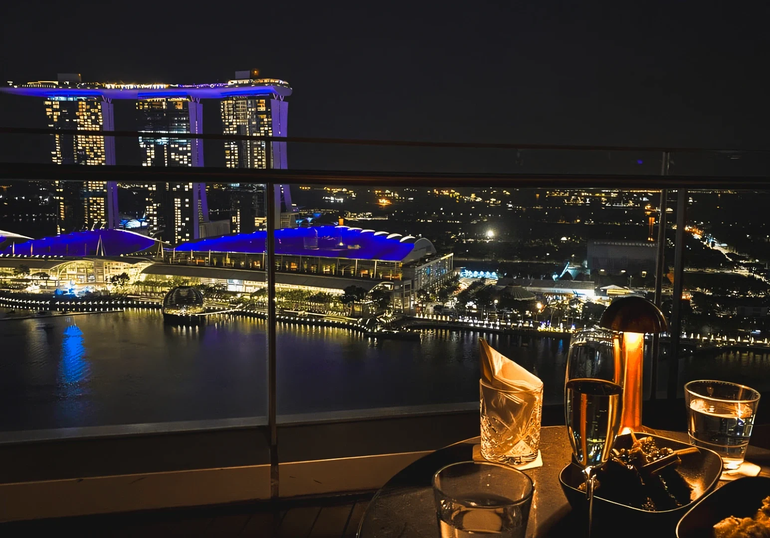2 Chasing After-Hours Bites High‑angle rooftop night shot overlooking a waterfront city skyline with illuminated landmarks, luxury buildings, and drinks on a table in the foreground, showcasing upscale nightlife, city views, and rooftop dining atmosphere.