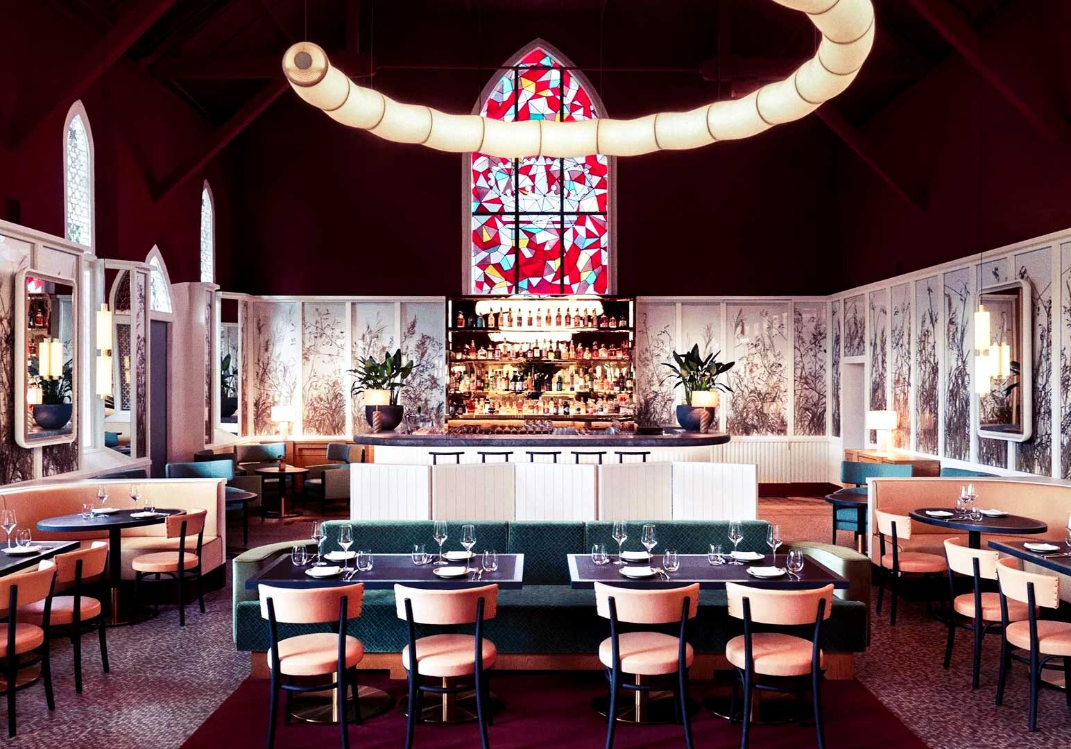 Elegant restaurant interior with a burgundy vaulted ceiling, serpentine pendant light, stained glass arched window, plush peach chairs, green banquettes, and a symmetrical backlit bar.