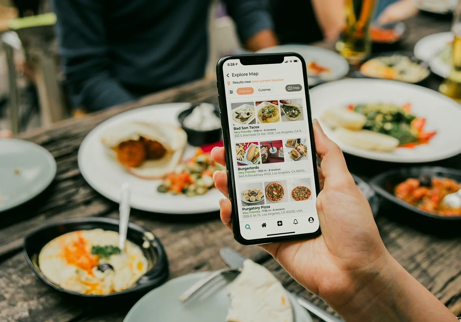 Eye-level close-up of a hand holding a smartphone displaying a food discovery app with restaurant deals, taken over a dining table filled with shared plates and drinks.