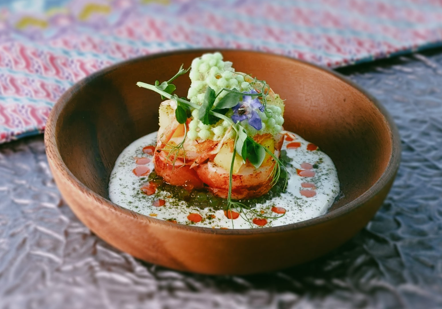 Gourmet seafood dish plated in a wooden bowl, featuring lobster pieces, microgreens, edible flowers, and a creamy sauce accented with herb oil and red droplets.