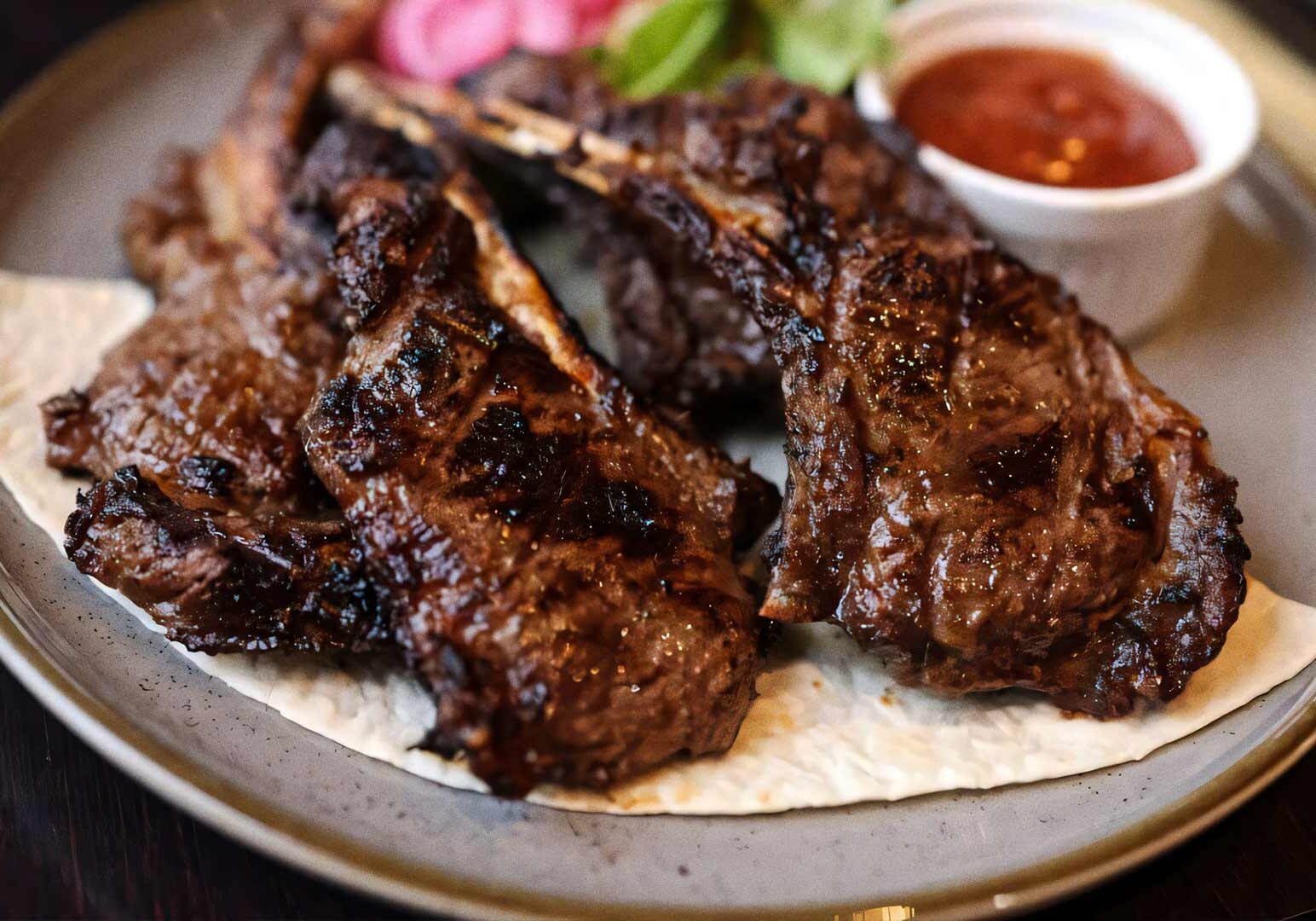 Grilled lamb chops served on flatbread with charred finish, red dipping sauce, pickled vegetables, and fresh greens garnish.