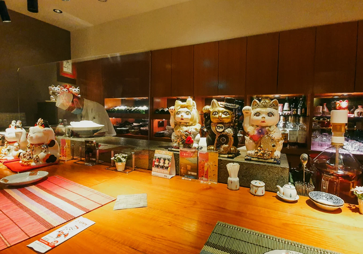 2 Hanashizuku Japanese Cuisine Warm wooden sushi counter at Hanashizuku Japanese Cuisine featuring maneki-neko figurines, sake bottles, and intimate dining ambiance ideal for Japanese food lovers, shot angle: eye-level wide interior perspective from seat at counter.