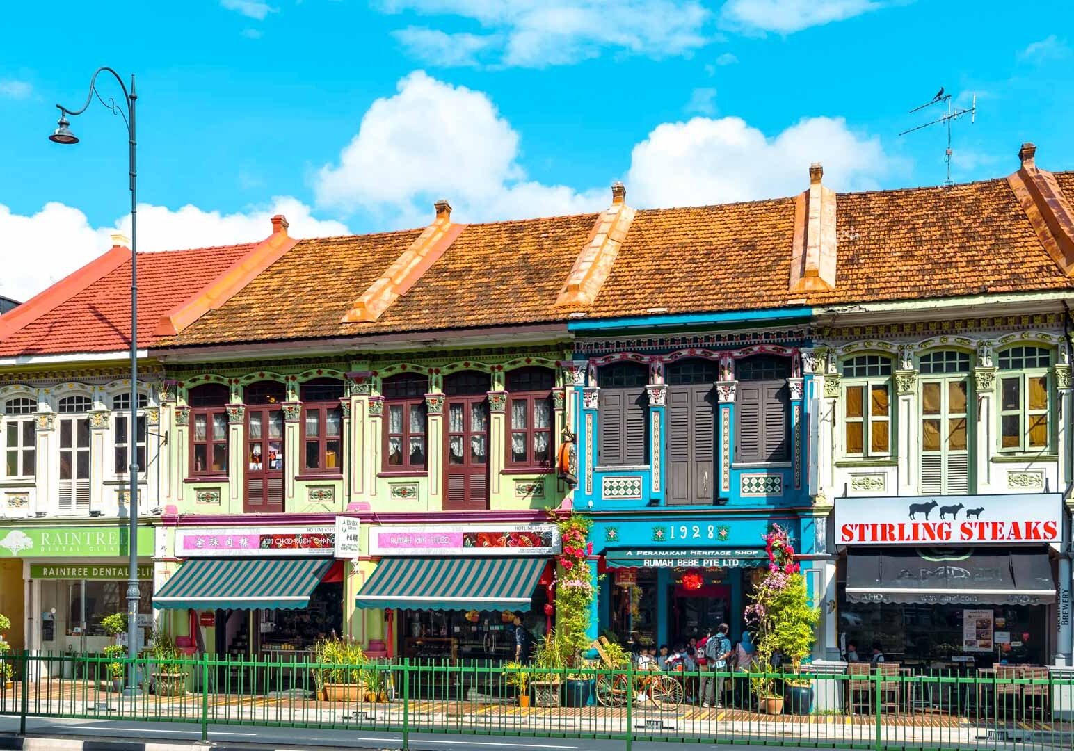 Row of colorful heritage shophouses with ornate windows and wooden shutters along a vibrant street, featuring cafes and shops under striped awnings on a sunny day.
