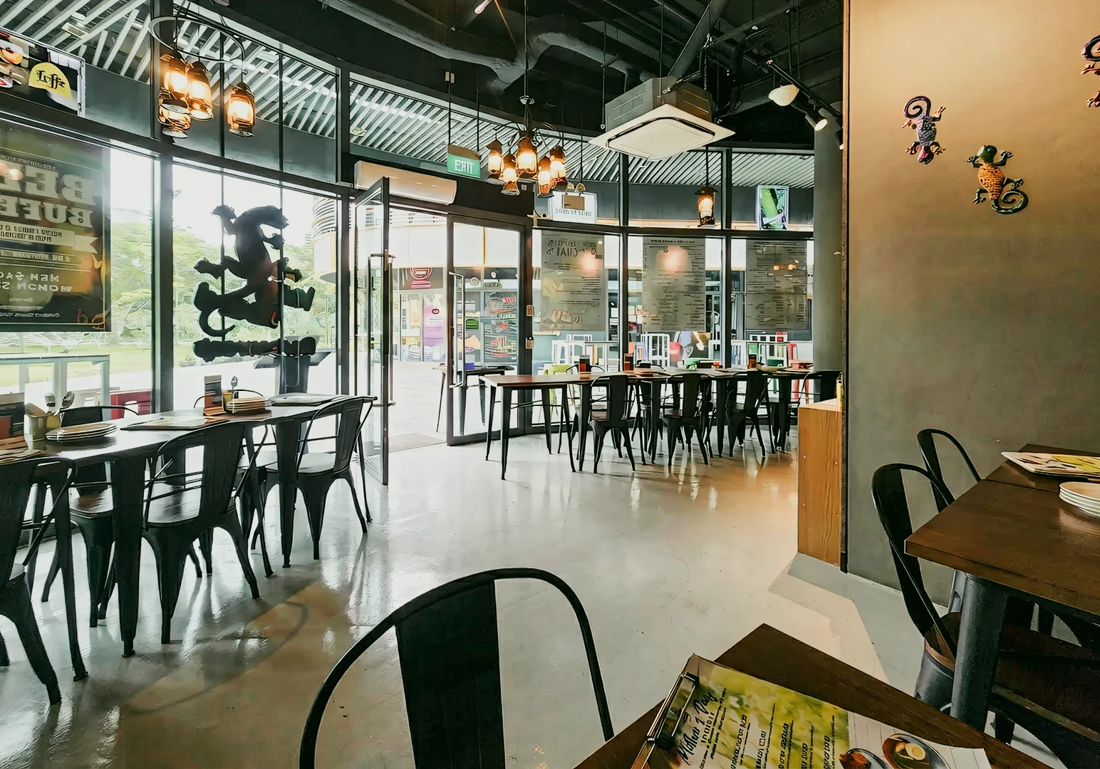 2 Lazy Lizard Flora Vista Wide-angle interior shot at eye level of a modern casual restaurant dining area with metal chairs, wooden tables, hanging pendant lights, and large glass windows.