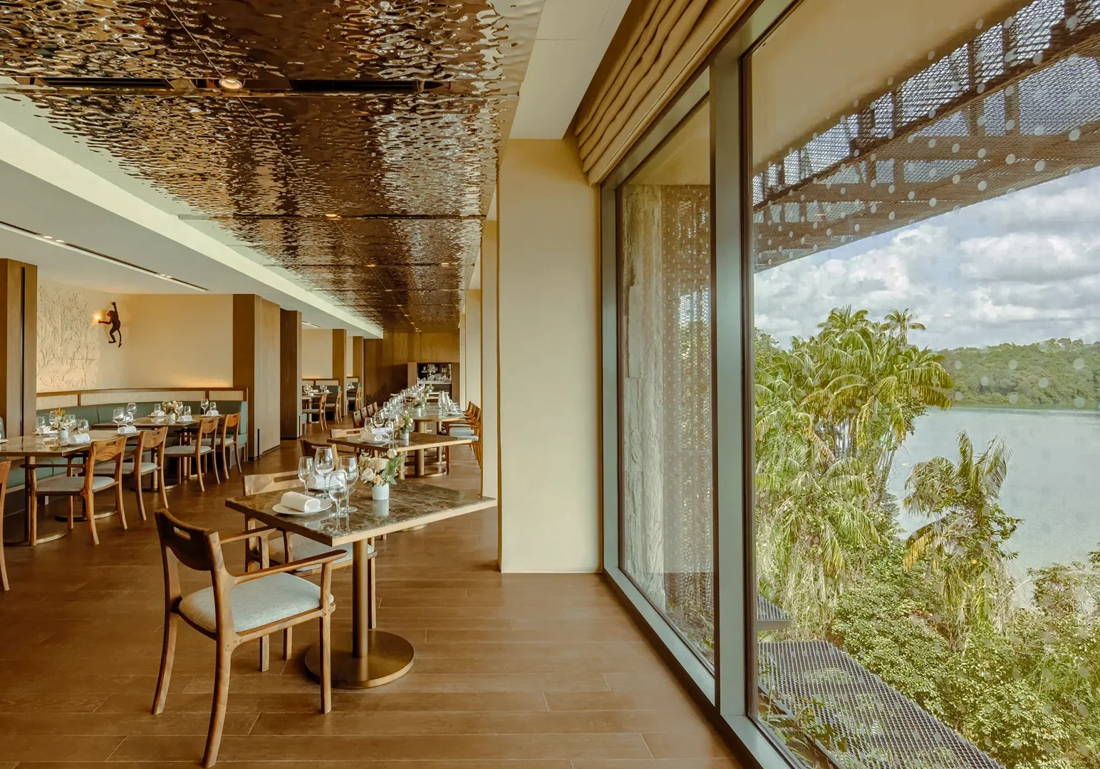 2 Mandai Rainforest Dining Wide‑angle eye‑level shot of an elegant rainforest‑inspired restaurant interior with neatly set dining tables, wooden flooring, textured ceiling panels, and floor‑to‑ceiling windows overlooking lush greenery and a water view.