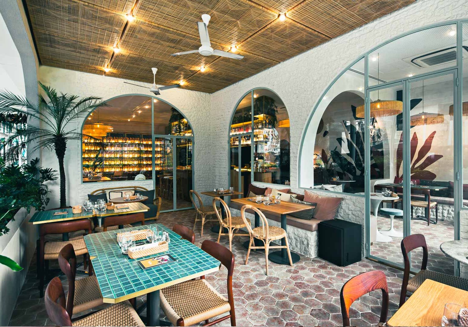 Interior of a stylish café featuring arched glass doorways, woven ceiling panels, wooden tables, and lush indoor plants creating a bright, modern dining space.