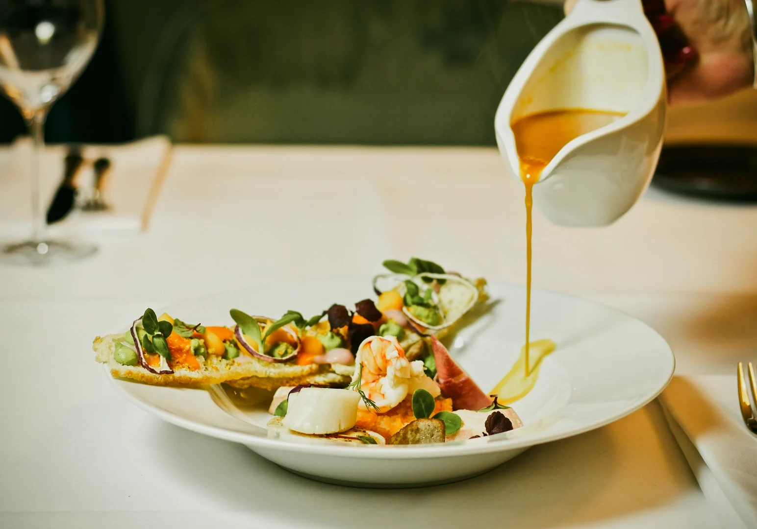 Close-up eye-level shot of a chef pouring sauce over a plated gourmet dish at a Michelin-starred restaurant, highlighting fine dining presentation.