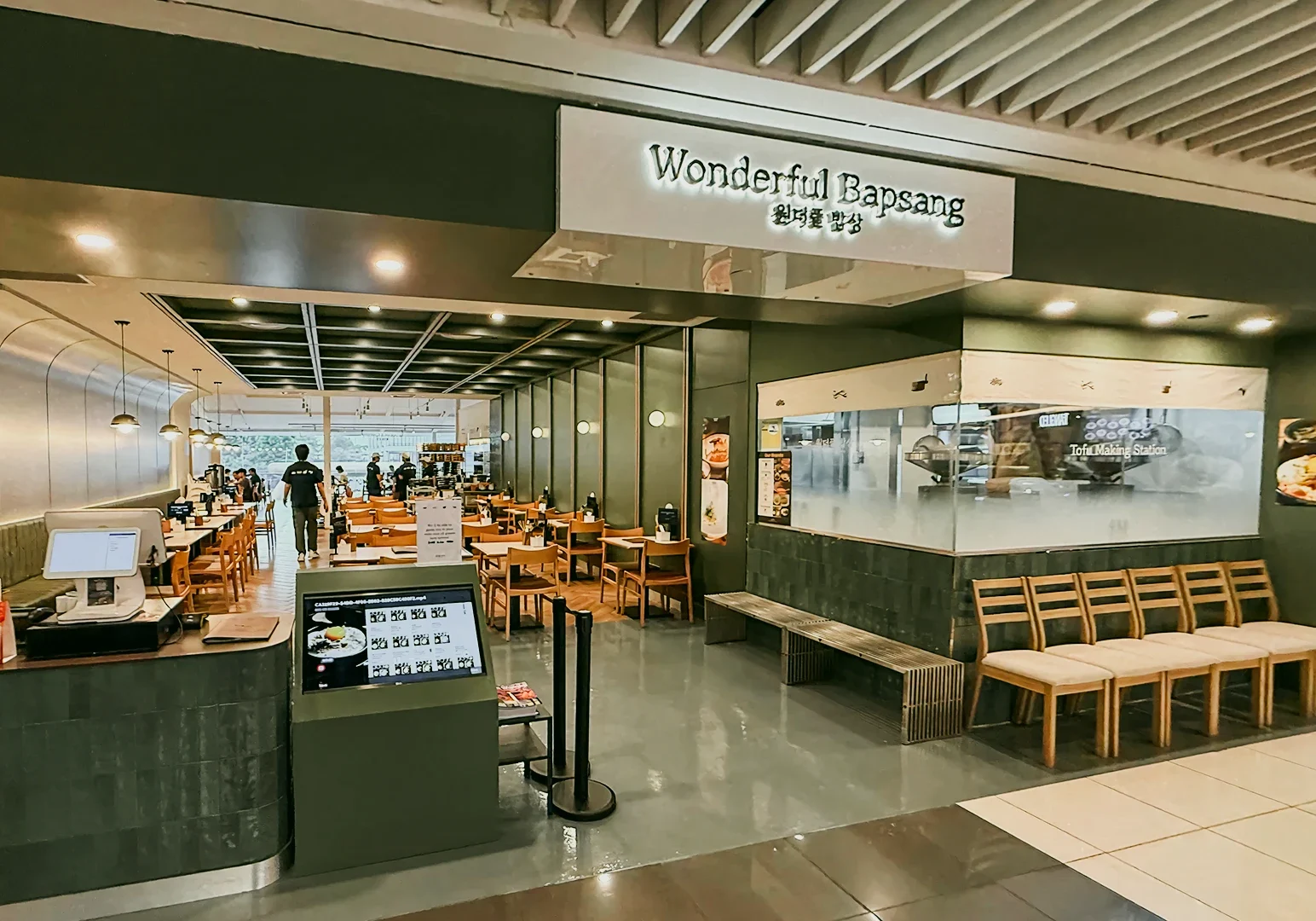 Straight-on medium-wide shot of the Wonderful Bapsang restaurant exterior in a shopping mall, showing empty dining tables, front counter, digital menu screen, and seating area.