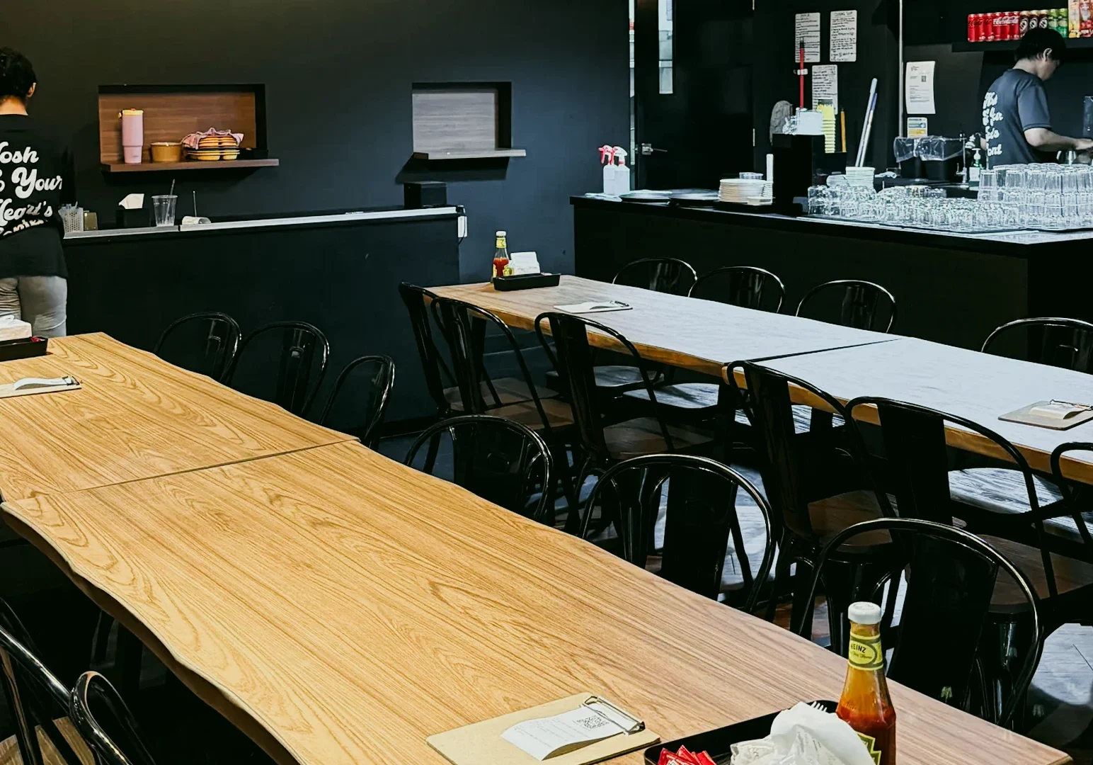 Wide-angle eye-level shot of a modern casual restaurant interior with long wooden tables, black chairs, open counter area, and minimalist industrial décor.