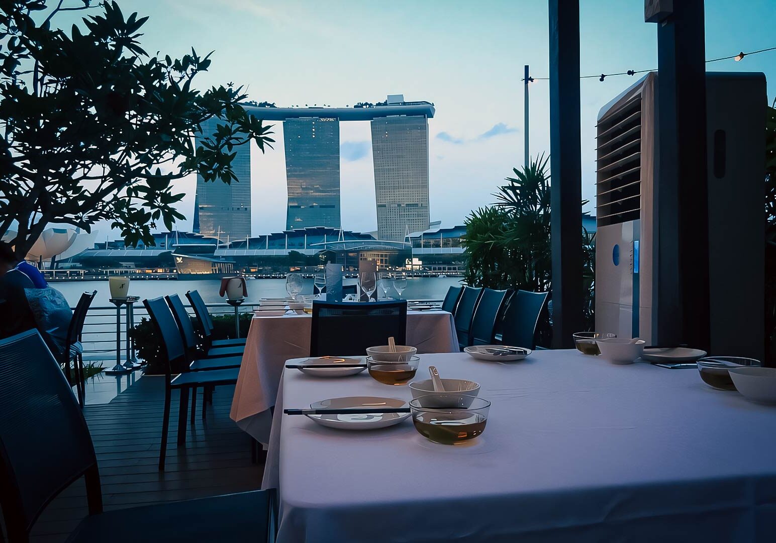 2-Palm-Beach-Seafood Outdoor waterfront dining setup overlooking Marina Bay Sands at dusk, featuring neatly arranged tables, white linens, and scenic views of Singapore’s skyline.