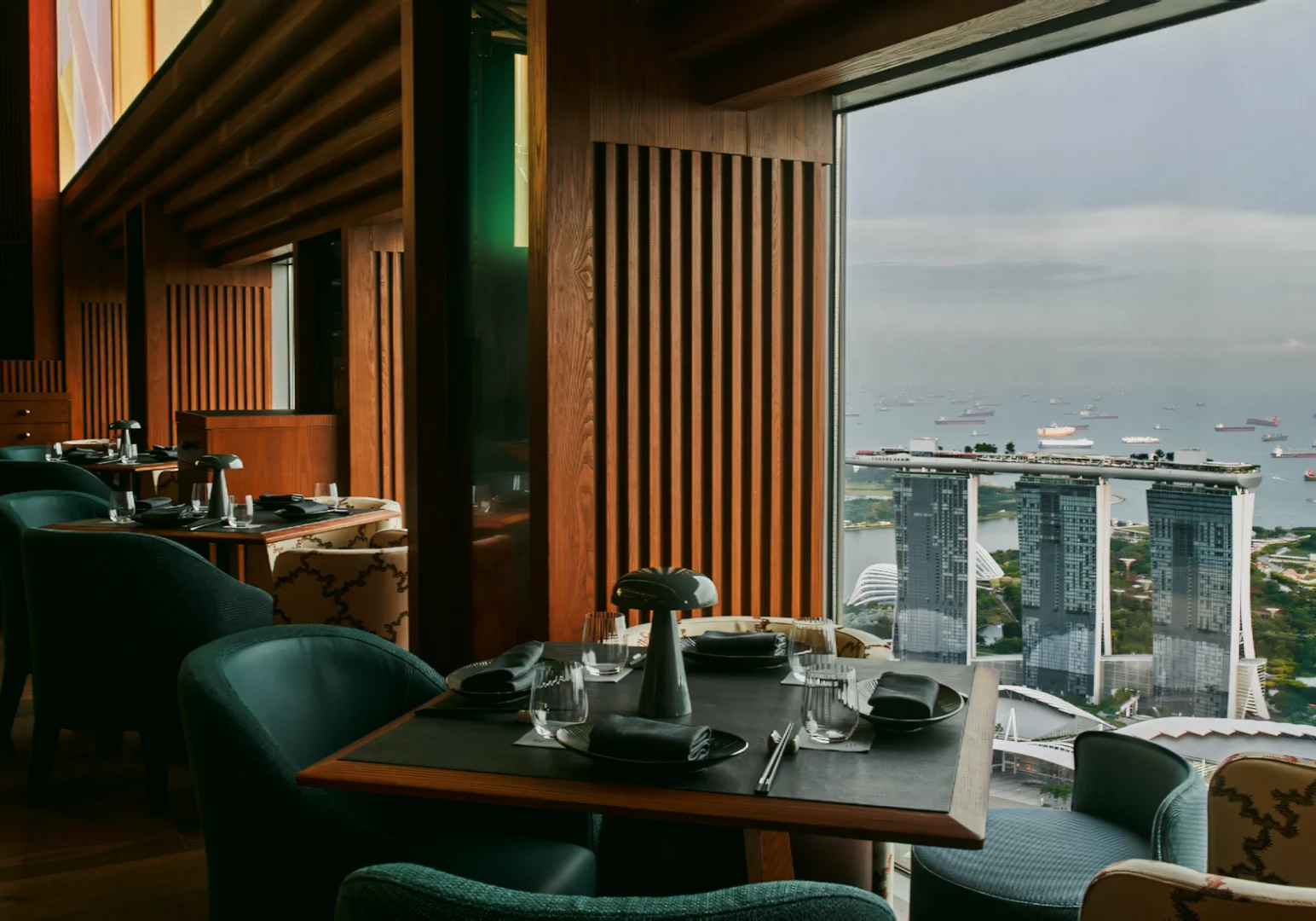 Eye‑level wide‑angle interior shot of a luxury restaurant dining table beside floor‑to‑ceiling windows overlooking Marina Bay Sands and Singapore harbor.