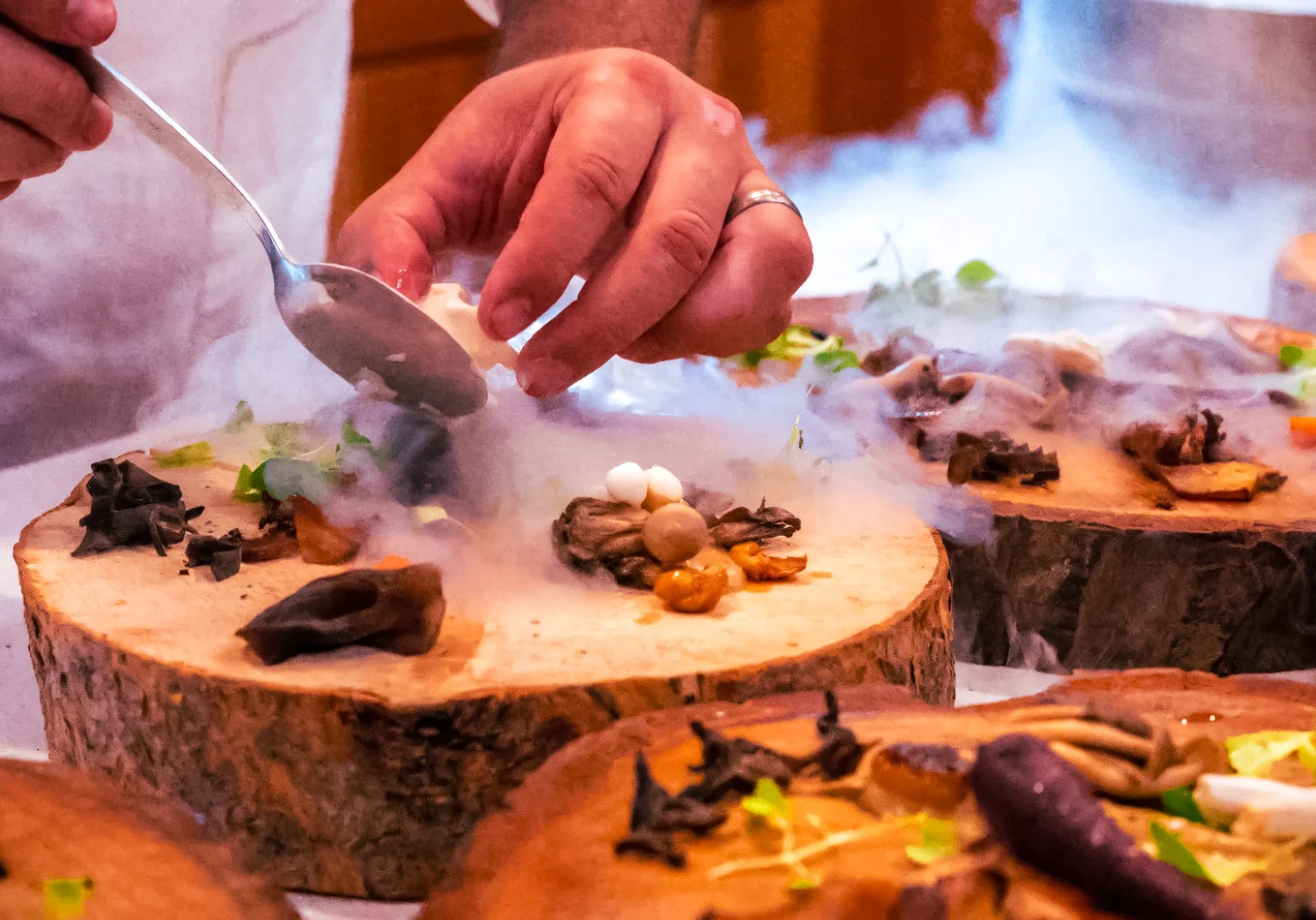 Close‑up shot of a chef’s hands carefully spooning mushrooms and herbs onto a wooden slab, with vapor rising from liquid nitrogen to create a smoky, dramatic effect.