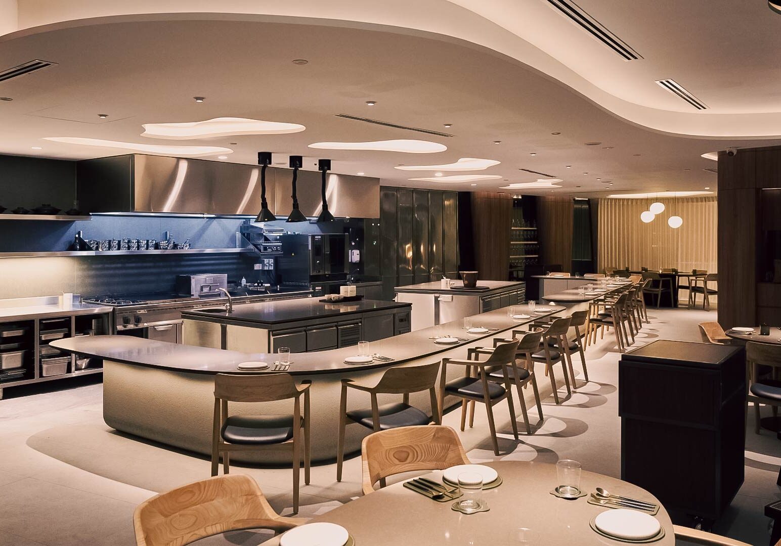 Contemporary fine‑dining restaurant interior with an open kitchen, curved countertops, minimalist wooden chairs, and warm ambient lighting.