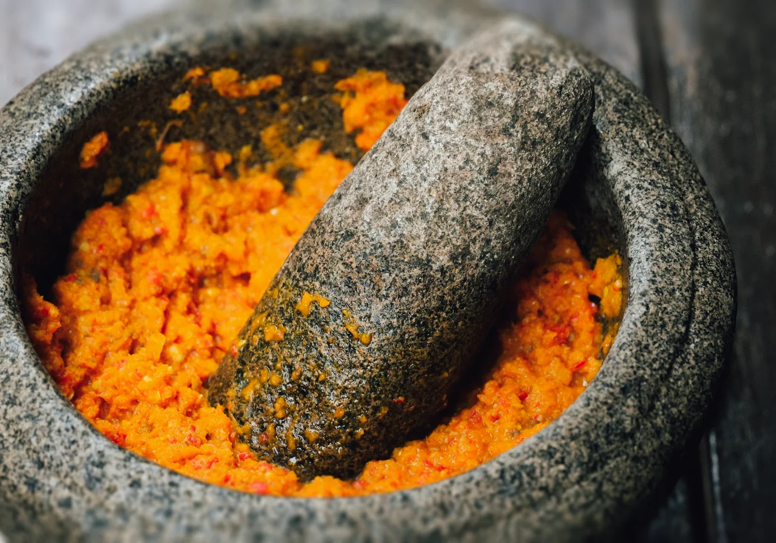 2 Singapore’s Culinary Past Restaurants Close‑up shot of rempah being ground in a stone mortar and pestle, showing a vibrant orange spice paste made from chilies and aromatics, highlighting traditional Peranakan and Southeast Asian cooking techniques and heritage cuisine preparation.