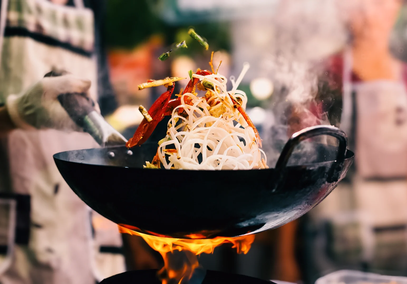 Street food vendor stir‑frying noodles with vegetables and seafood in a flaming wok, showcasing fresh ingredients, high heat cooking, and authentic Asian street food preparation.