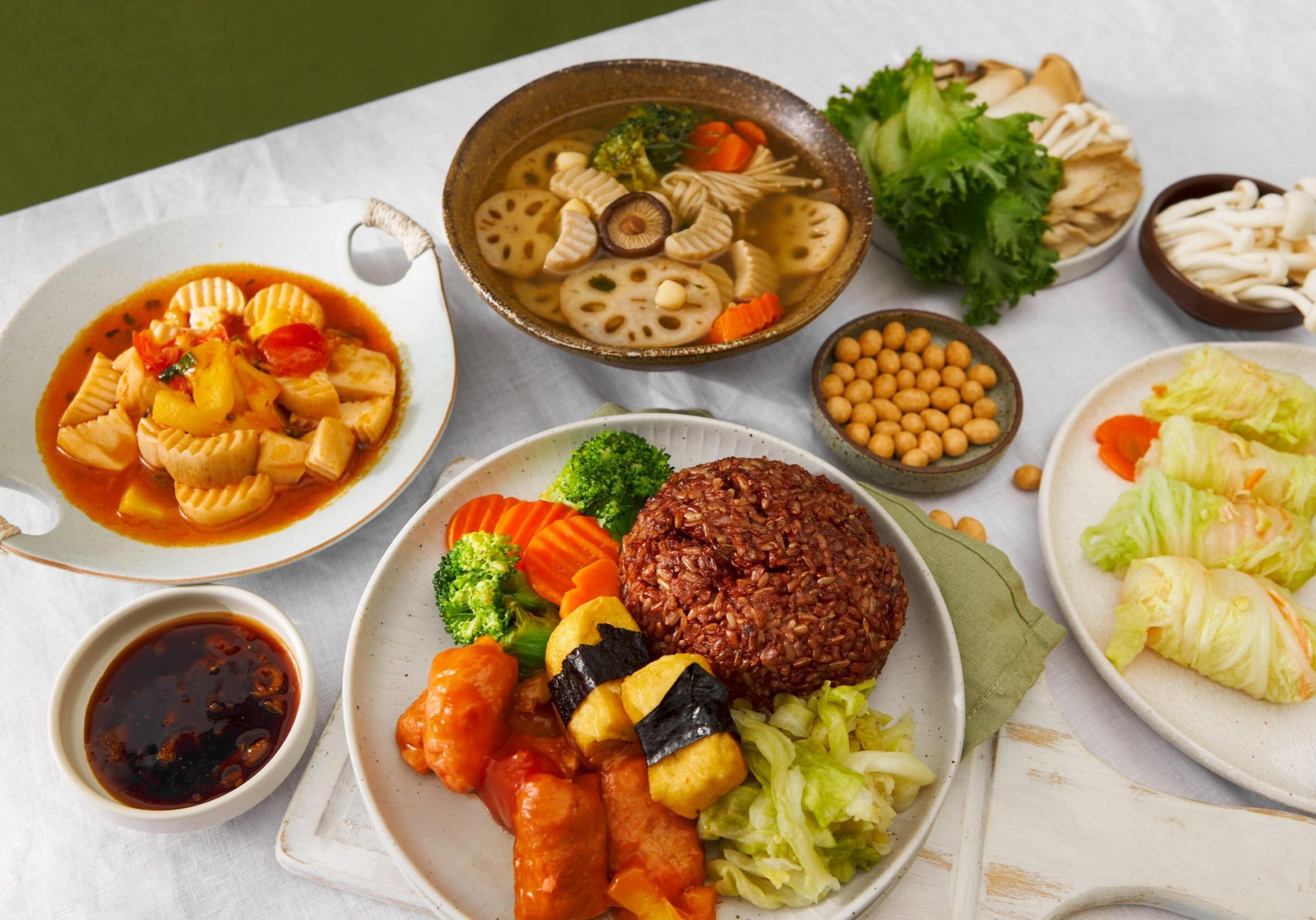 This image showcases a vibrant spread of vegetarian dishes, including a plate of red rice served with steamed broccoli, carrots, and tofu wrapped in seaweed. Surrounding the main plate are various sides such as a clear vegetable soup with lotus root, cabbage rolls, and a bowl of fresh mushrooms and greens.
