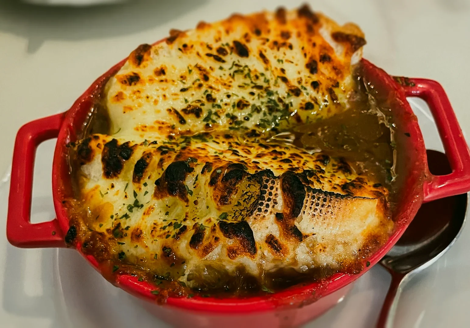2 The Secret Garden By Zeekri Top-down close-up shot of French onion soup served in a red ceramic crock, topped with toasted baguette slices and bubbling melted cheese, showing rich broth and classic bistro presentation.