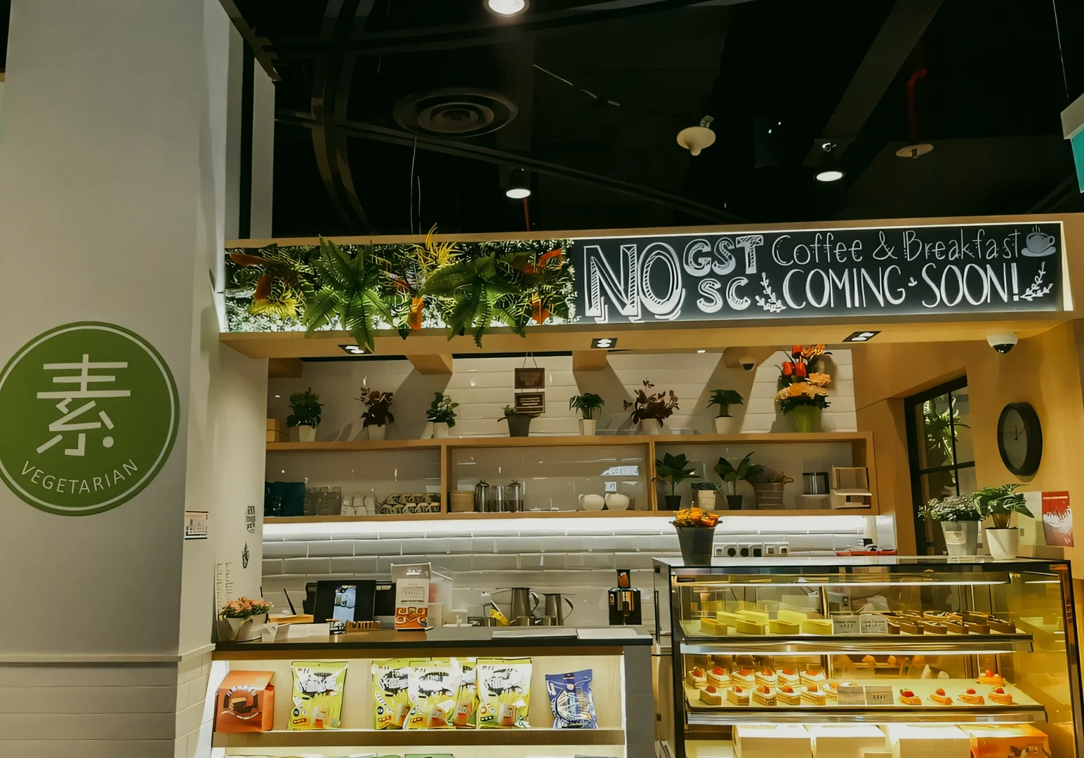 Eye‑level wide shot of a vegetarian café interior with a service counter, pastry display case, hanging plants, and visible signage indicating a vegetarian and coffee breakfast concept.