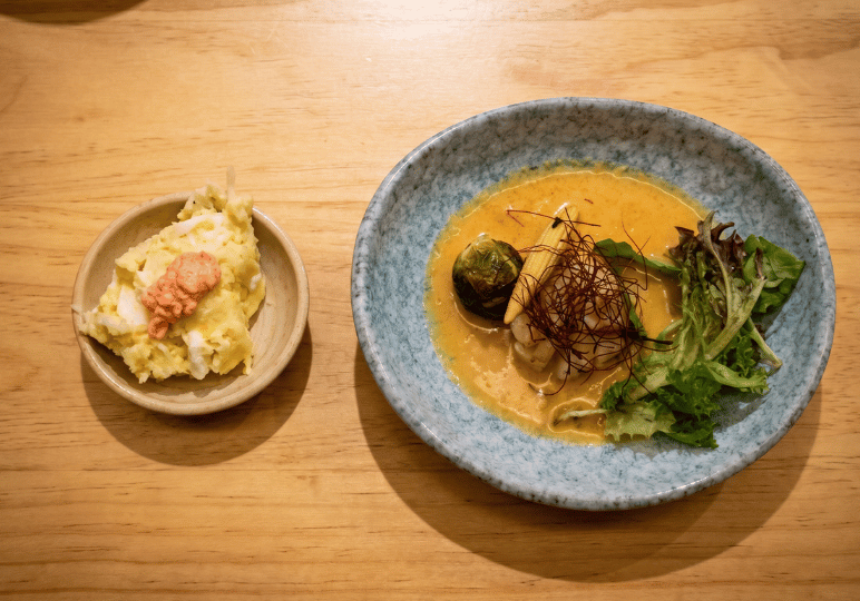 A top-down view of two dishes on a wooden table: a small bowl containing potato salad topped with mentaiko, alongside a larger blue ceramic bowl holding a fish fillet in a rich yellow curry sauce with greens.