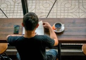 2-clarissa-chua-Writers High-angle view of a man in a black t-shirt reading a manuscript at a long wooden desk with a cup of black coffee and a smartphone.