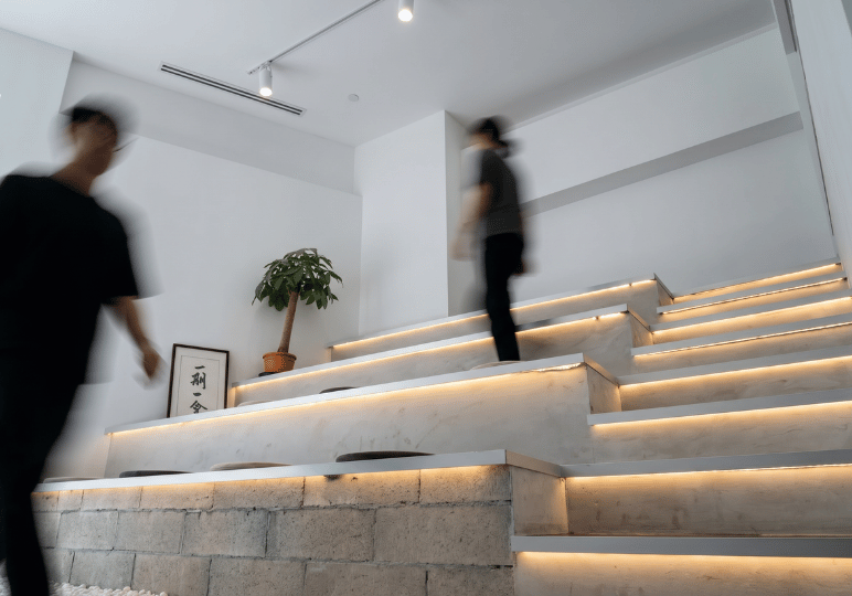 Blurred figures in motion walking up a tiered, stadium-style seating area made of white stone with wooden edges, warm under-lighting, and flat seat cushions.