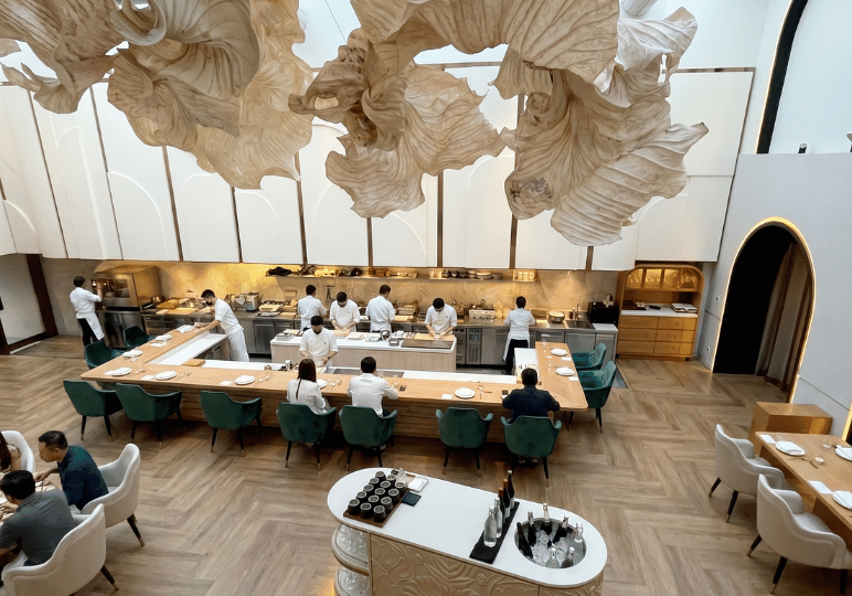 2-restaurant-interior A bright, modern dining room with a high ceiling, featuring a large curved wooden chef's counter, plush green dining chairs, and dramatic, oversized leaf-like sculptural installations hanging from above.