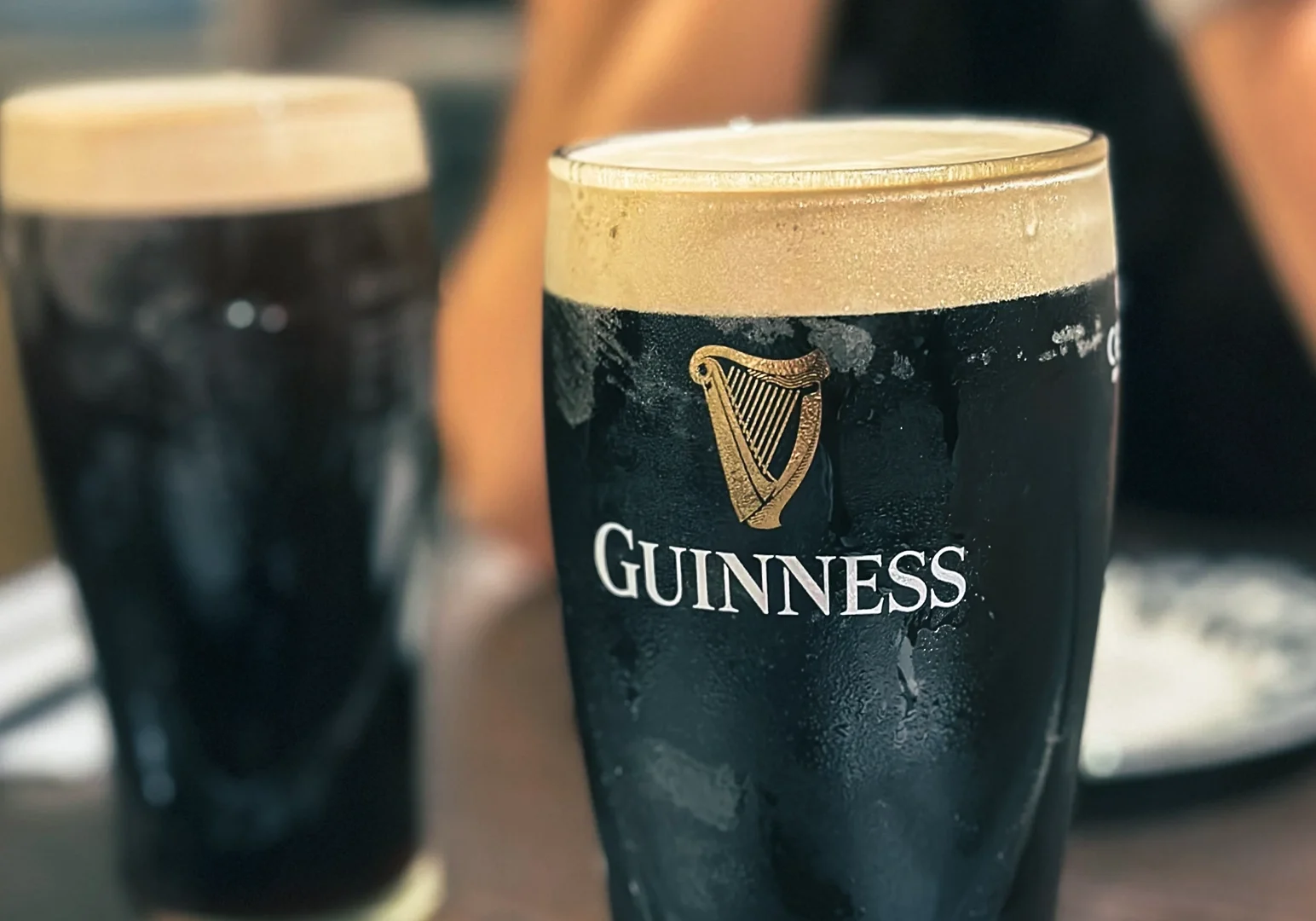 Close-up of a chilled pint of Guinness stout with a creamy head, displayed on a table beside another glass of dark beer.