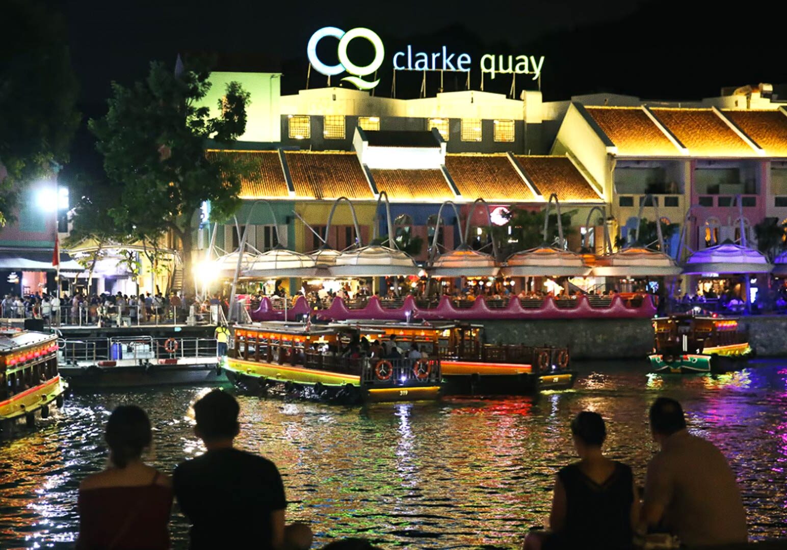 Nighttime view of Clarke Quay with vibrant lights and river cruise boats.