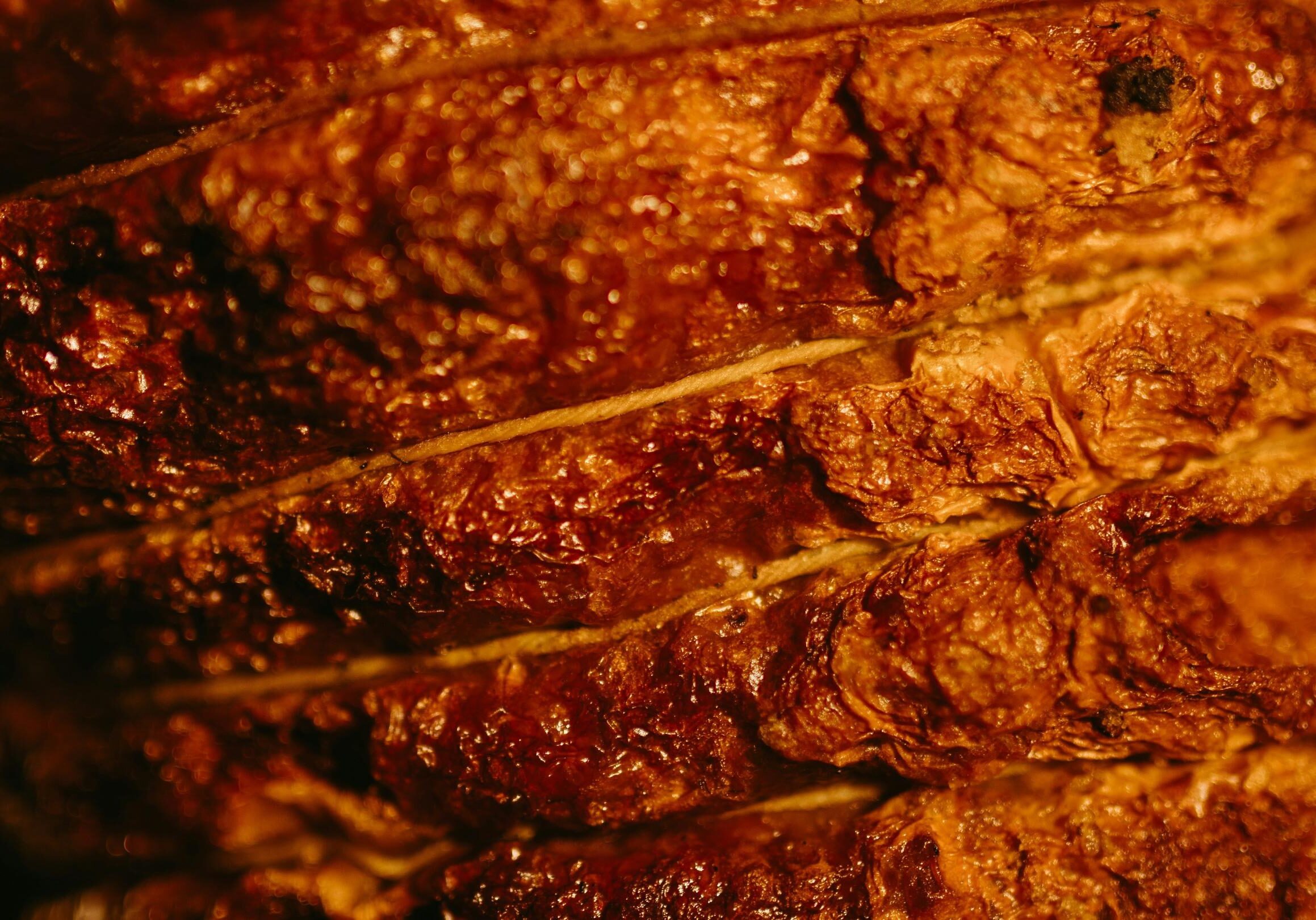 This extreme close-up features the deeply browned, textured surface of a roasted meat, possibly a brisket or prime rib, showing visible layers and rendering fat. The warm, amber lighting highlights the craggy, caramelized crust and the intricate details of the seared exterior.