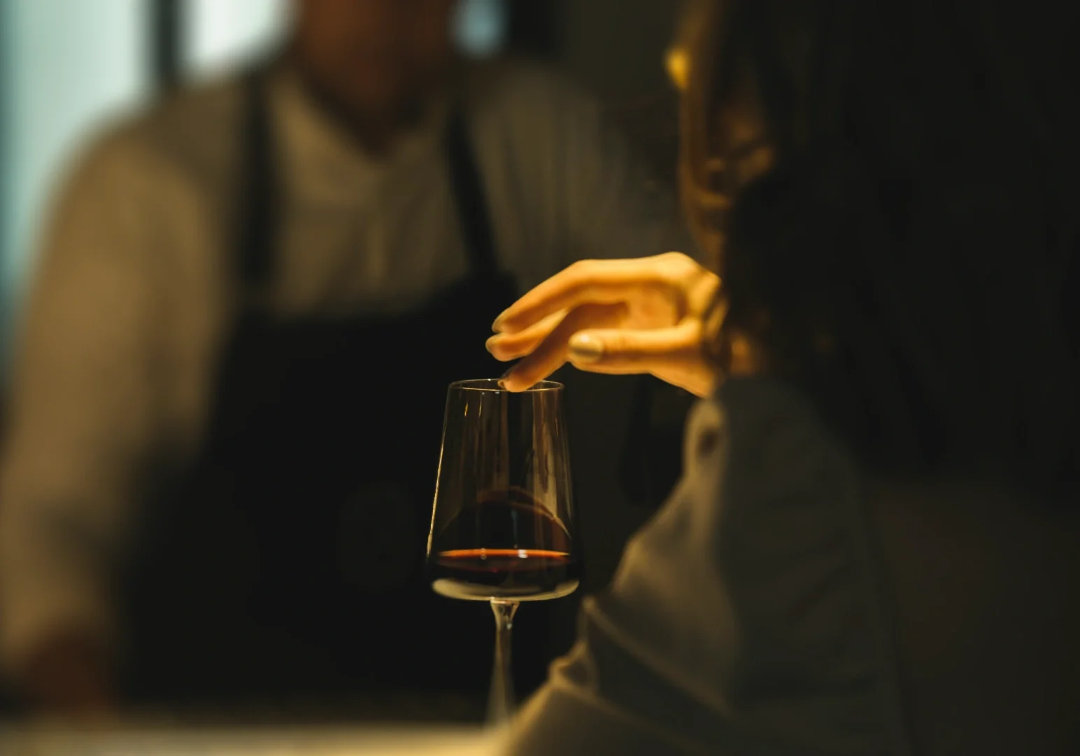 3 Chasing After-Hours Bites Shallow depth‑of‑field close‑up shot of a hand gently holding a glass of red wine at a dimly lit bar, highlighting refined nightlife ambiance, modern dining experience, and intimate after‑hours social moments.