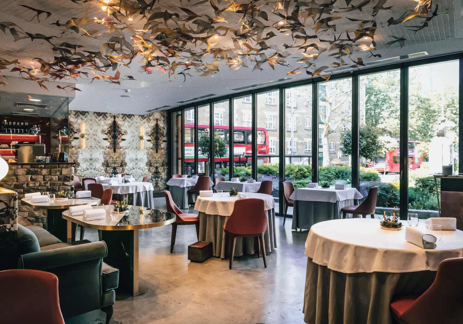 Upscale restaurant interior with round table setups, floor‑to‑ceiling windows, artistic ceiling decor, and views of passing red London buses outside.