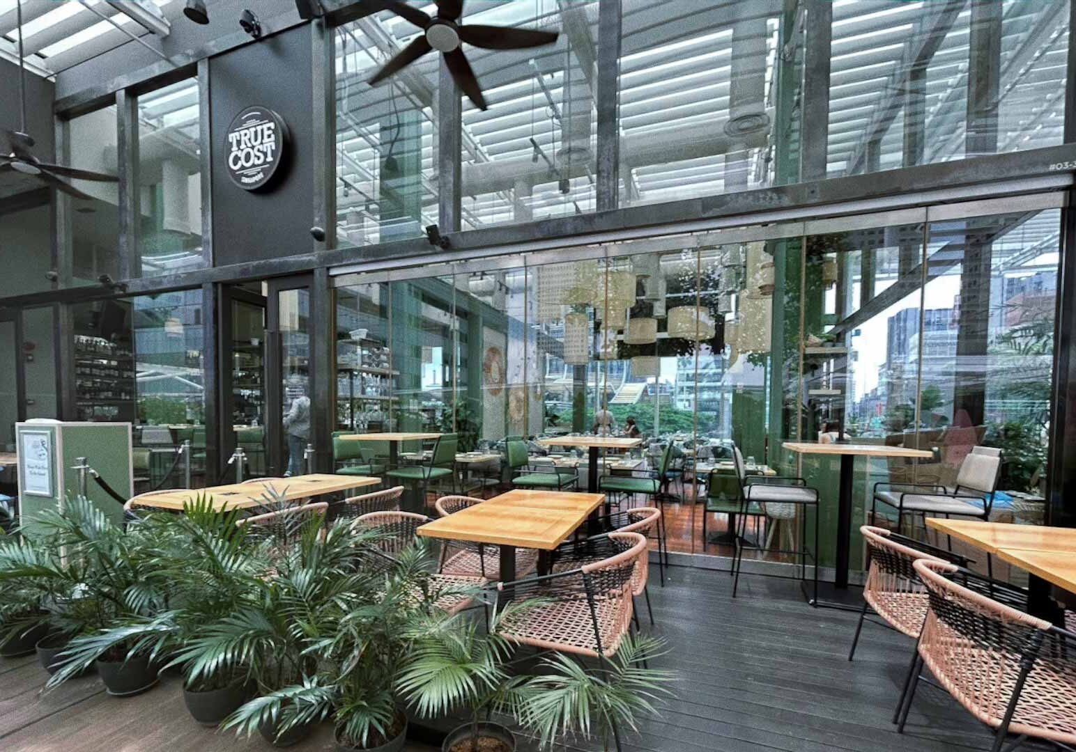 Outdoor dining area of the True Cost restaurant, featuring wooden tables, woven chairs, and potted plants along the perimeter.