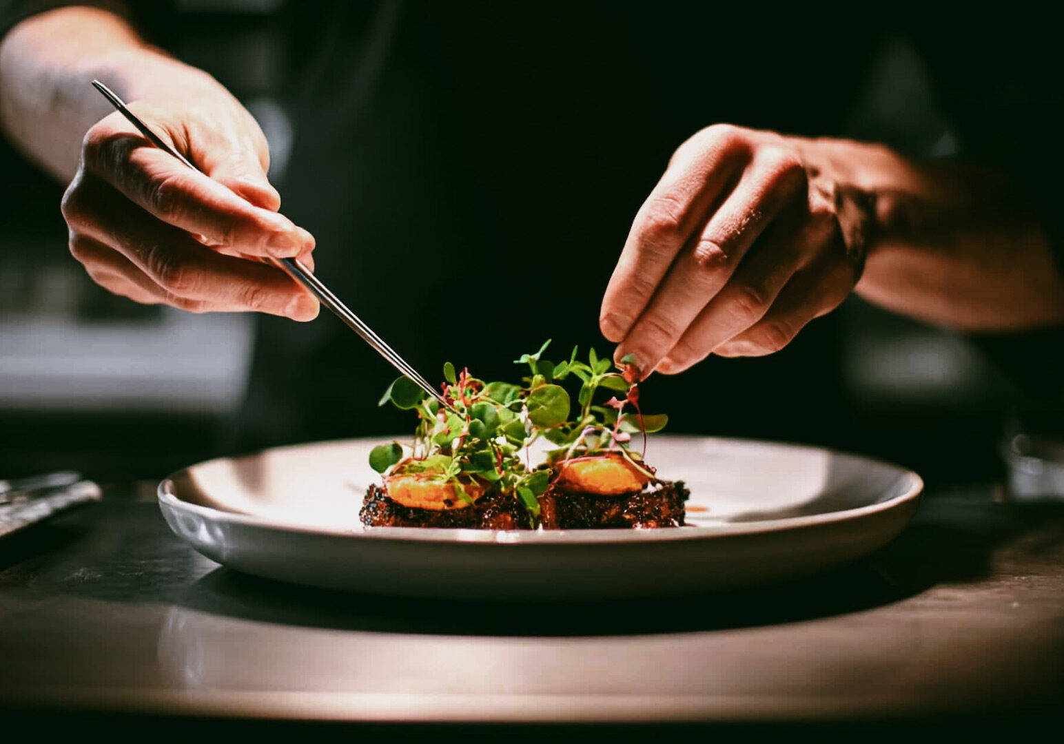 Chef carefully plating a fine‑dining dish using tweezers, arranging microgreens and vegetables on a white plate under focused kitchen lighting.