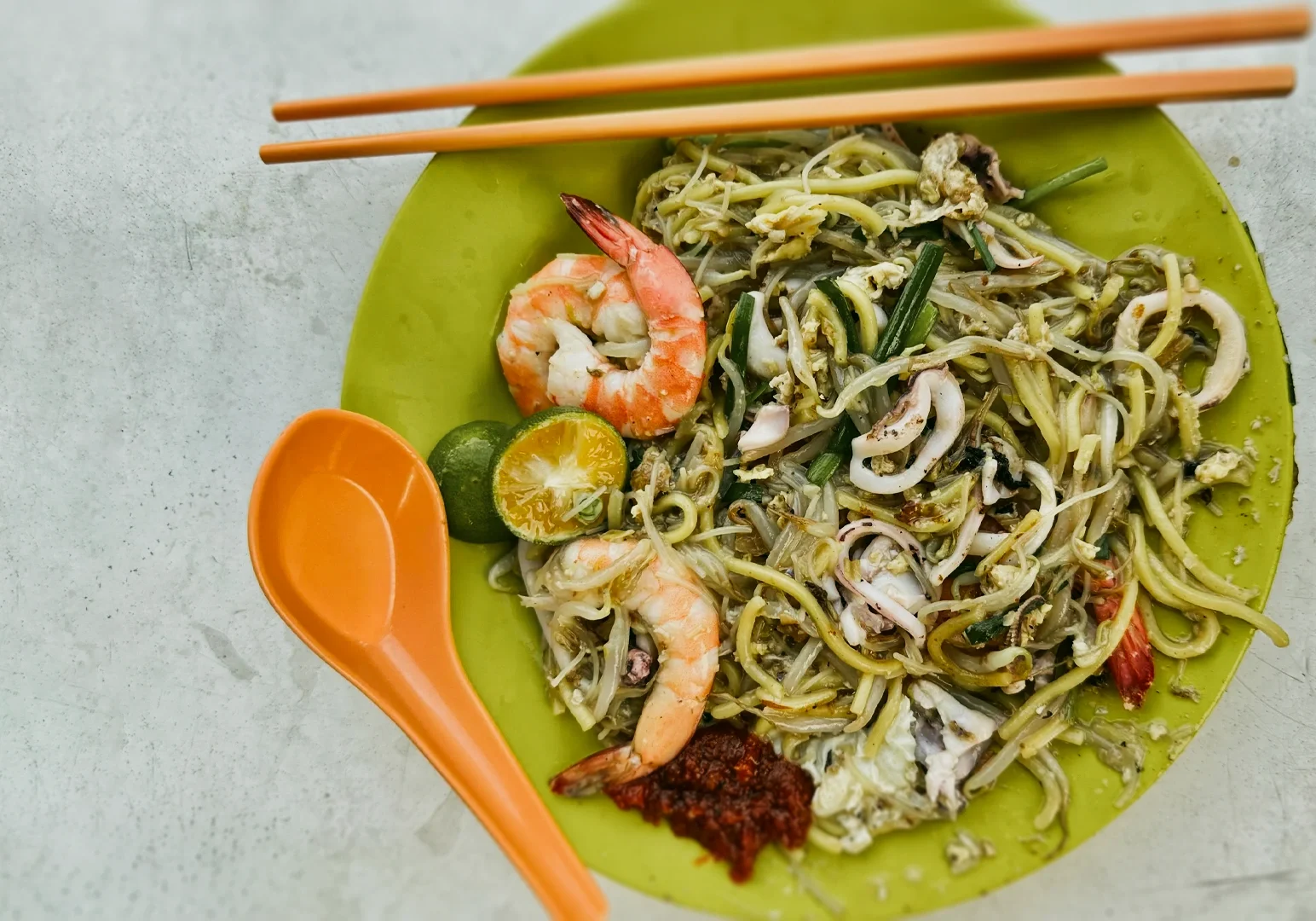 Singapore hawker-style Hokkien mee with prawns, squid, lime and sambal chili on a green plate, shot at a top-down overhead flat lay angle.