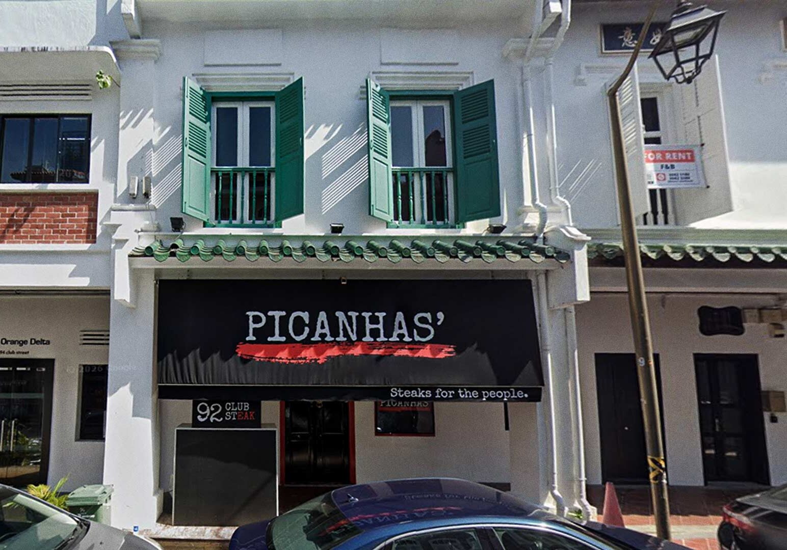 Picanhas’ steakhouse storefront featuring bold black awning with red underline, slogan ‘Steaks for the people,’ traditional architecture with tiled roof and green shutters, set among vibrant Club Street businesses.