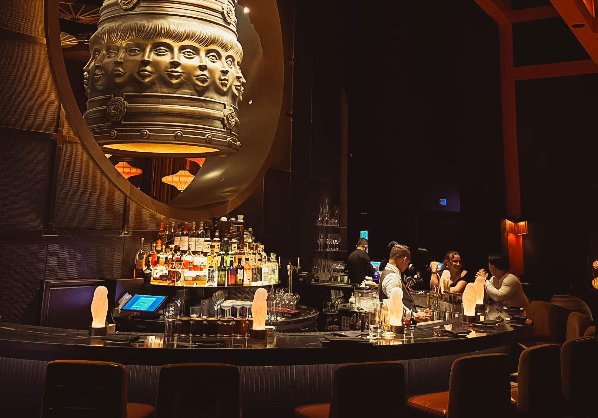Modern luxury bar interior featuring a large sculptural bronze bell centerpiece and professional bartenders serving cocktails.