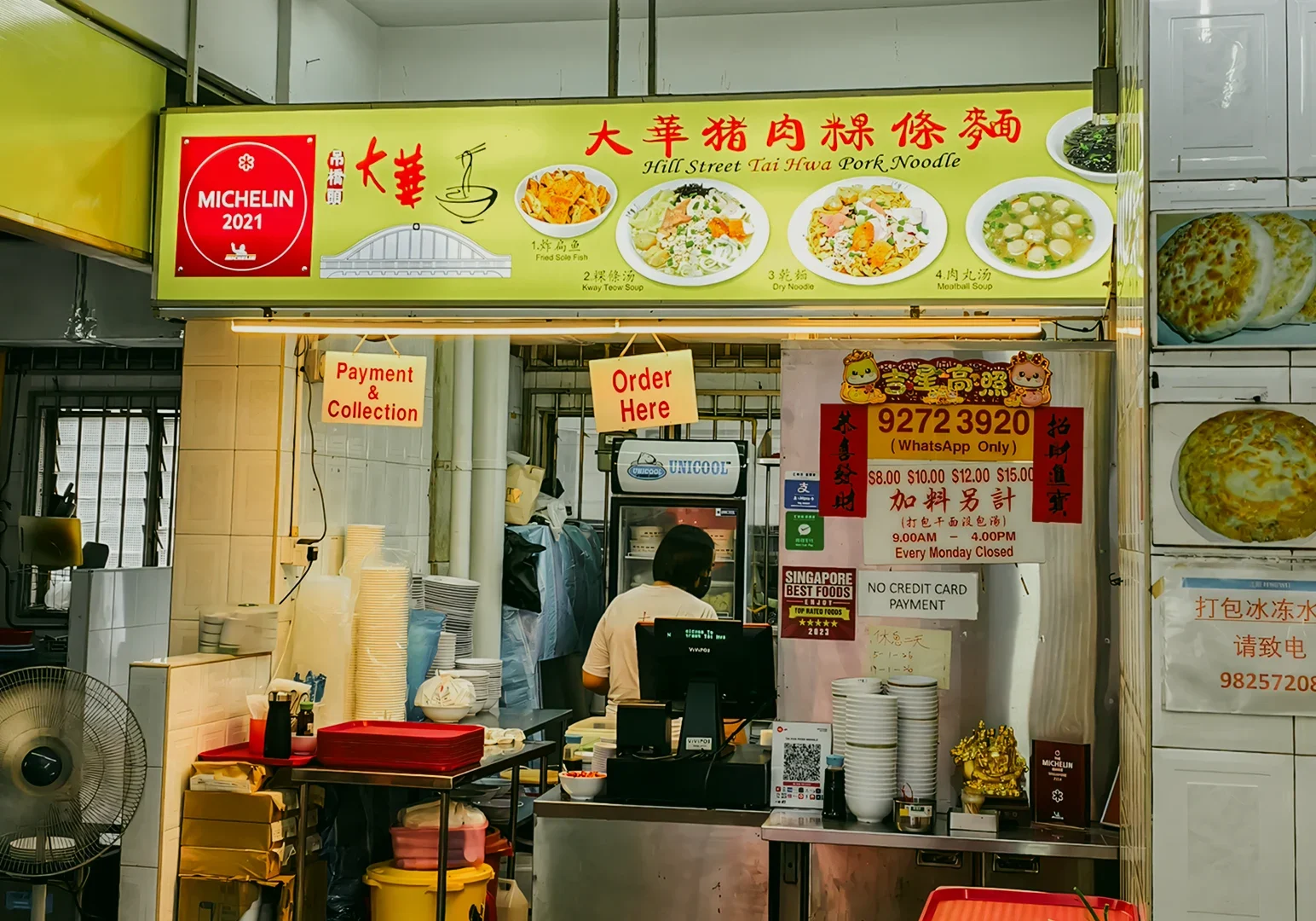 Eye-level wide shot of a bustling street food stall displaying a Michelin Guide sign, menu boards, and a vendor preparing noodles.