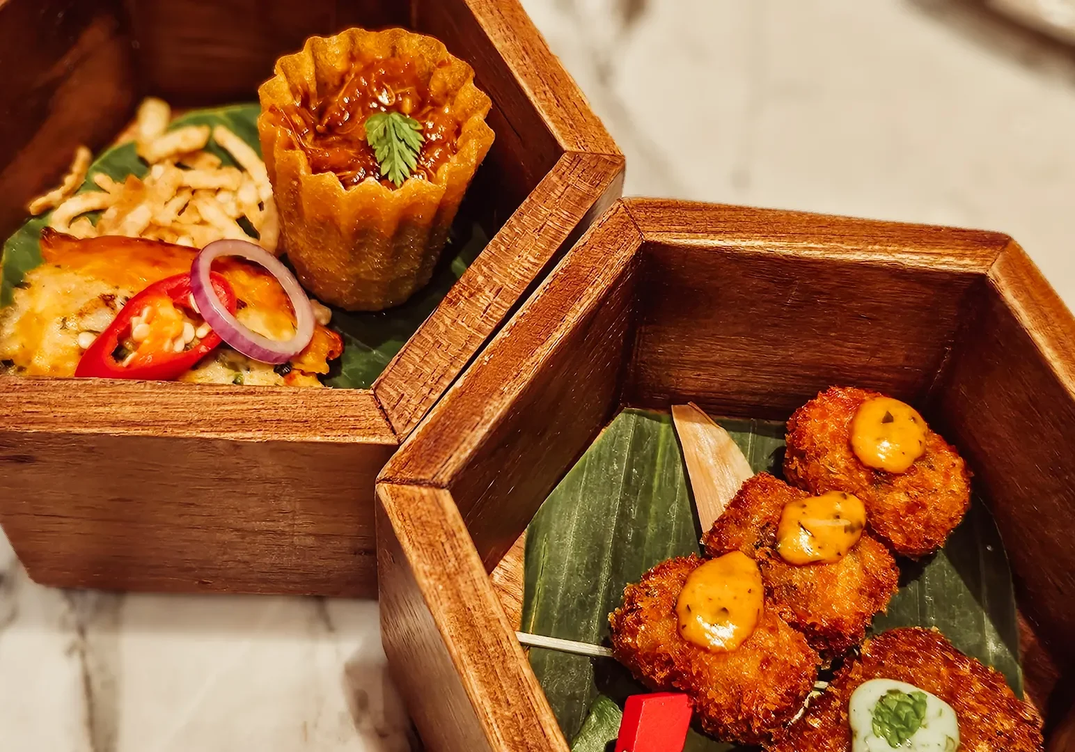 3 Jiak Kim House Singapore Wooden serving boxes filled with Peranakan-style appetizers, including kueh pie tee, fried skewered bites with spicy mayo, and a savory baked dish topped with sliced chili and onions.