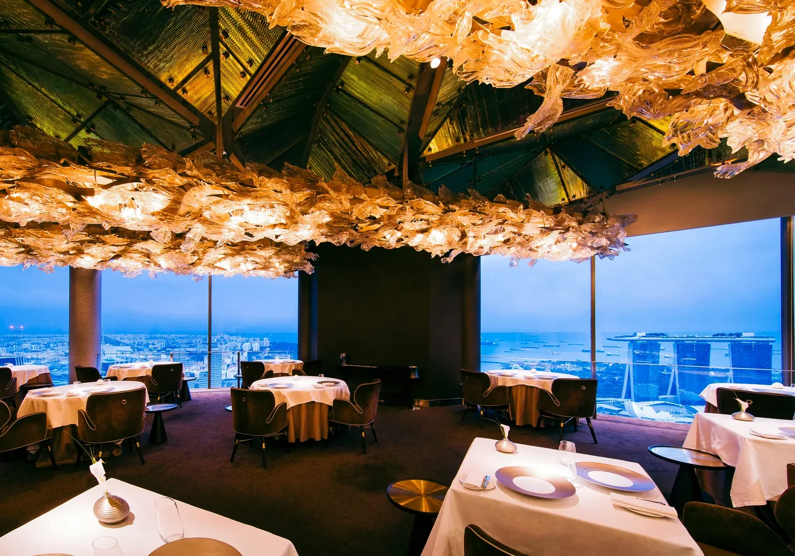 3 Kirk Westaway Upscale fine‑dining restaurant JAAN by Kirk Westaway featuring dramatic sculptural ceiling décor, round tables with white linens, and panoramic windows overlooking a city and waterfront skyline.