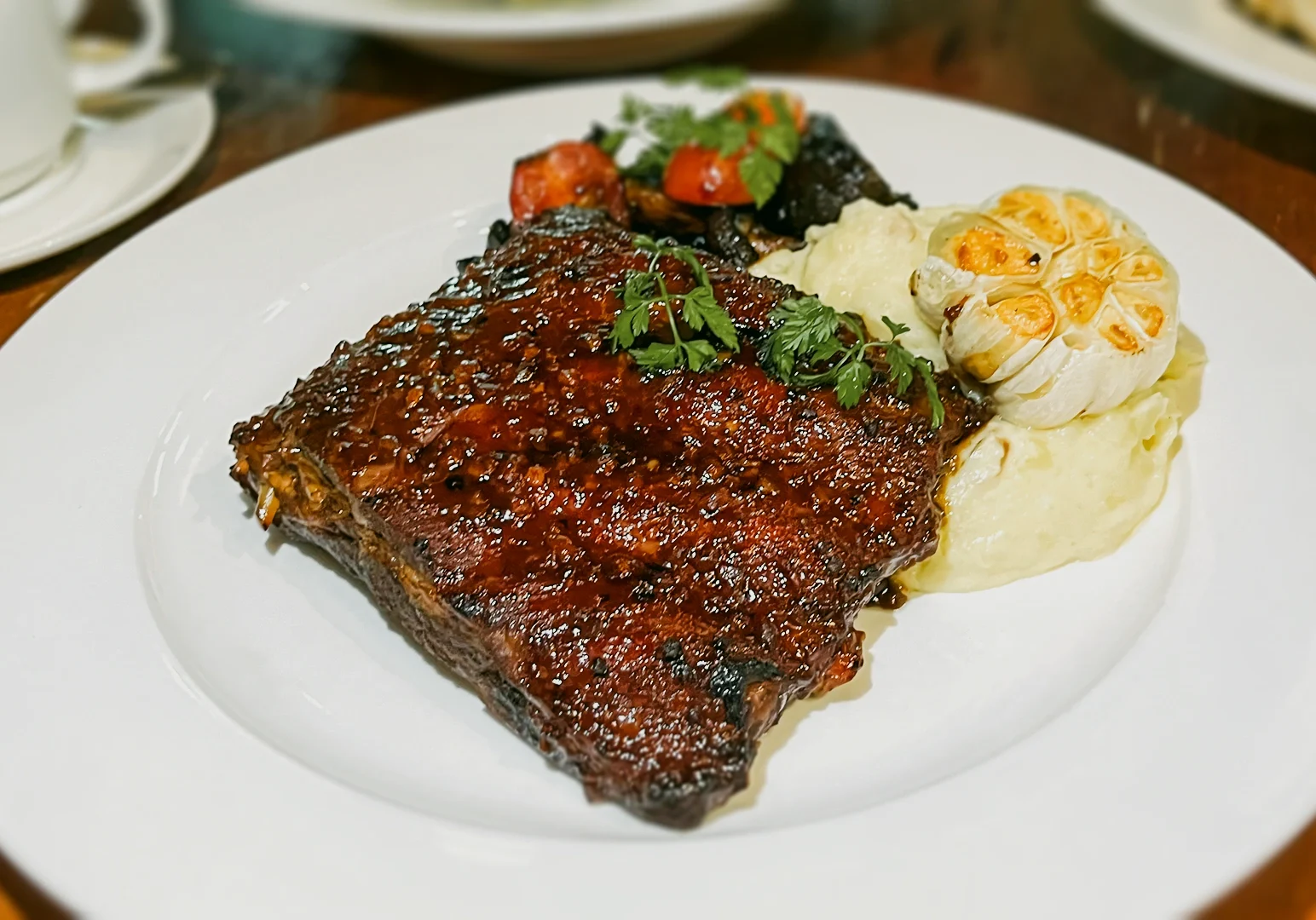 3 Lazy Lizard Flora Vista Close-up top-down angled shot of glazed barbecue back ribs, served with creamy mashed potatoes, roasted garlic, and vegetables on a white plate.