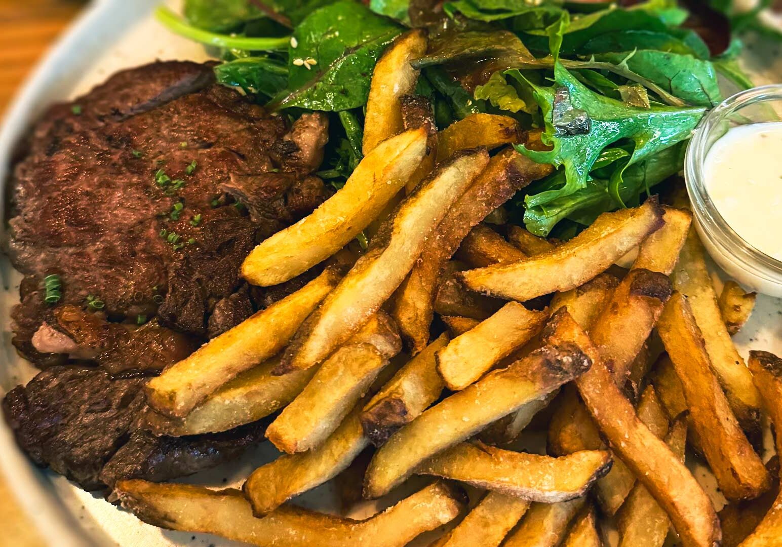 Plate of steak served with golden fries, mixed greens, and a small dish of dipping sauce on a rustic dining table.