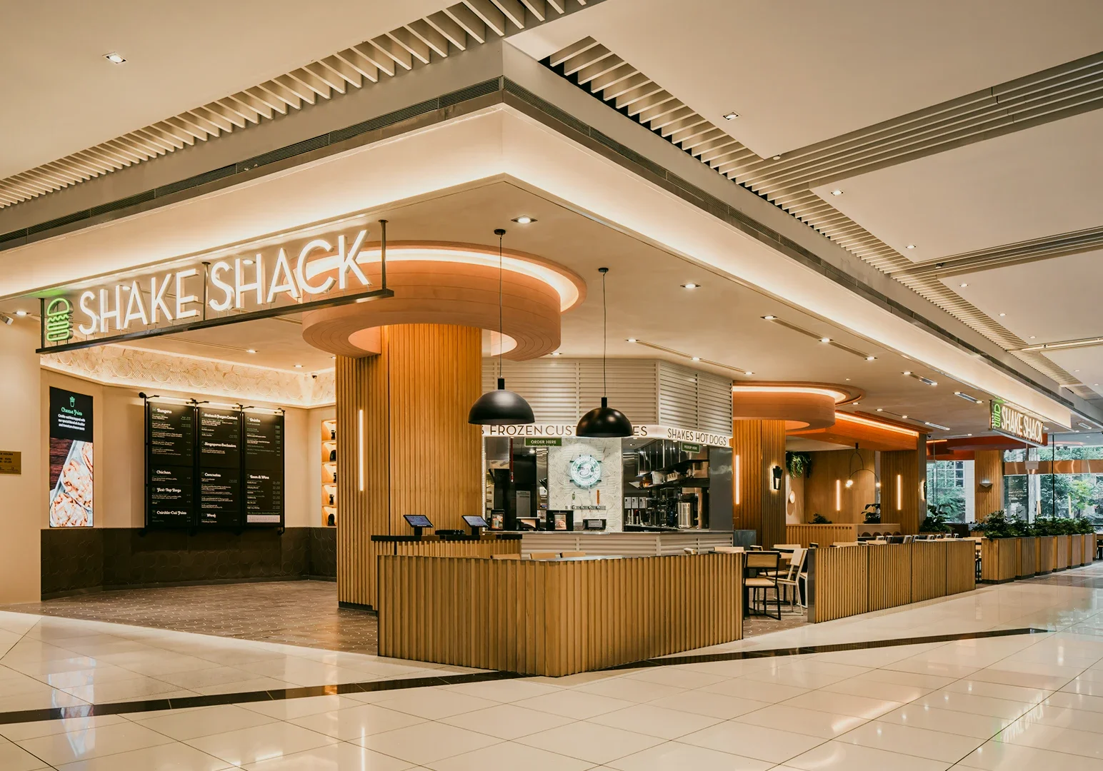 Wide-angle eye-level shot of a Shake Shack restaurant inside a modern shopping mall, featuring wood-accent interiors, ordering counters, seating areas, and bright overhead lighting.