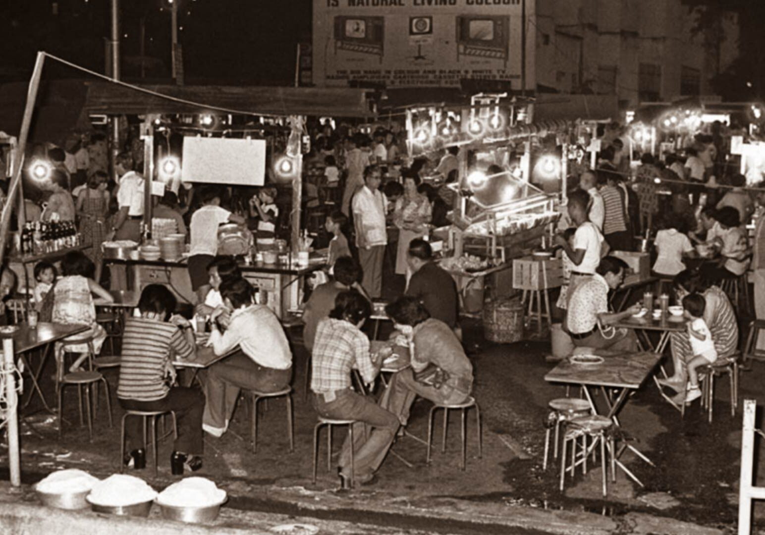 3-Palm-Beach-Seafood Historic black‑and‑white photo of a bustling Singapore street food scene at night, with hawkers cooking and diners seated at small stools and tables.