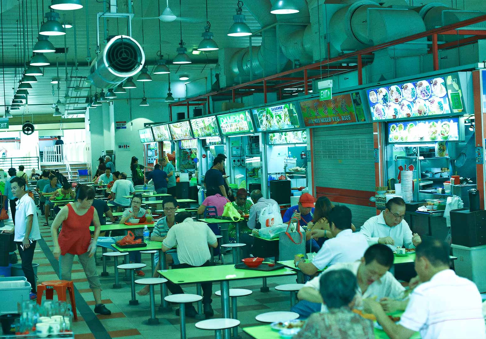 3-Tanjong-Pagar Bustling Tanjong Pagar hawker centre with green communal tables, busy food stalls, bright signboards, and diners enjoying local dishes.