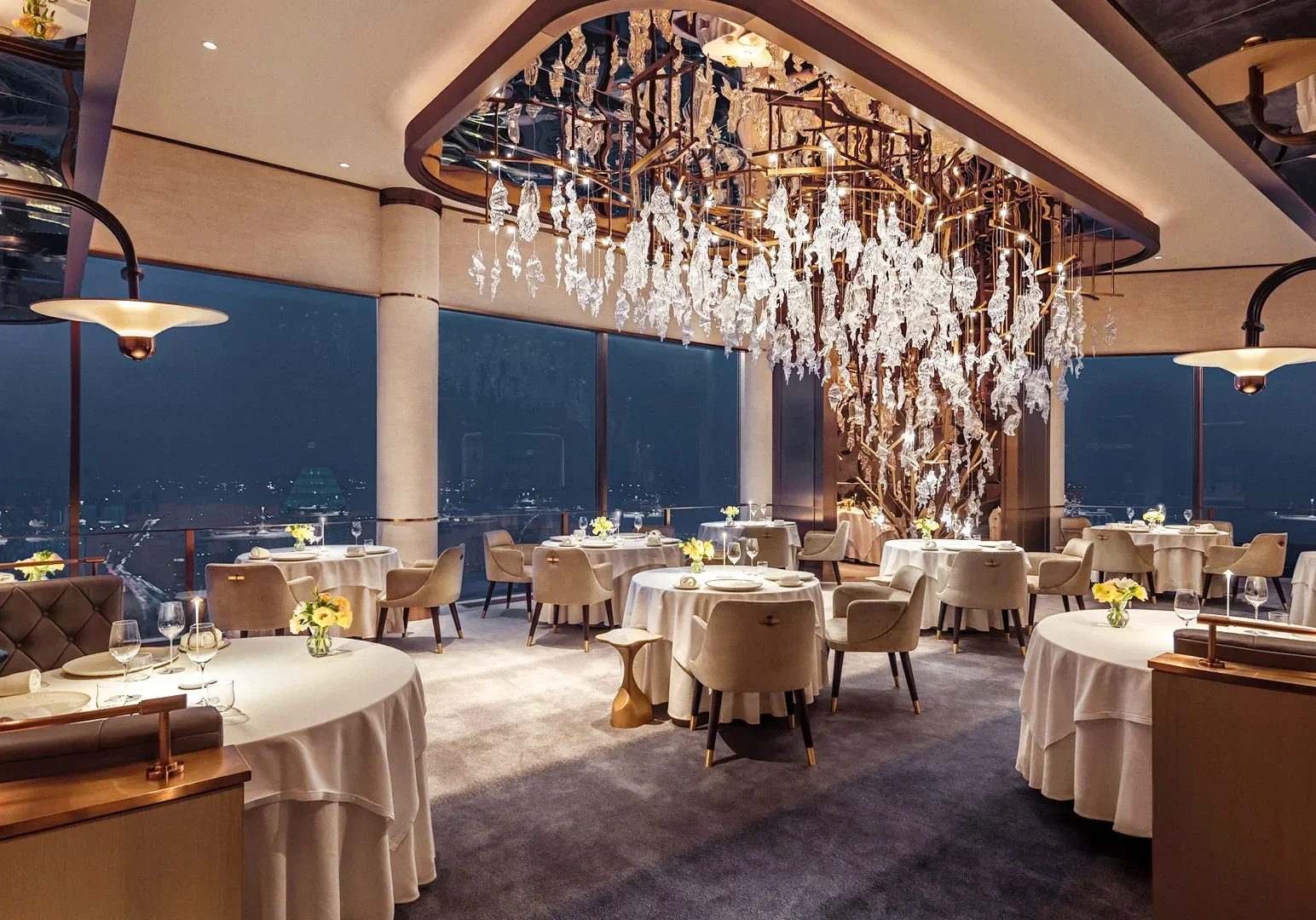 Elegant fine‑dining restaurant interior featuring round tables with white linens, floor‑to‑ceiling windows, and a sculptural crystal chandelier centerpiece.