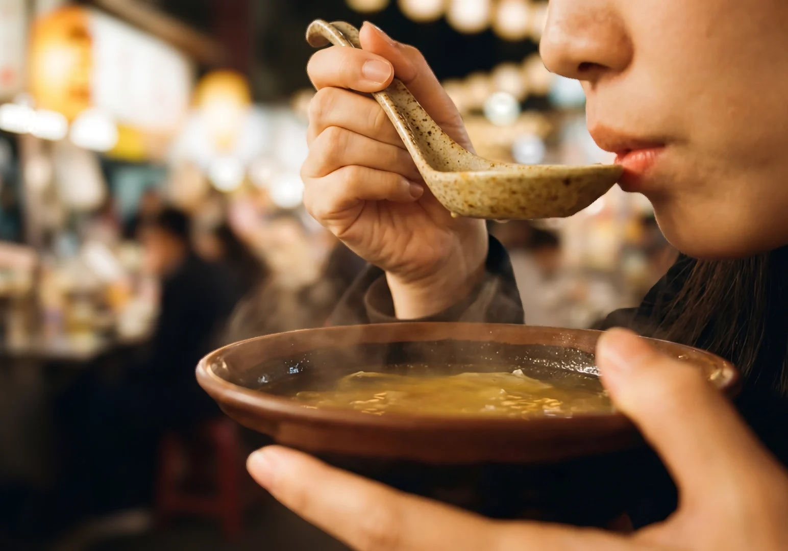 Close-up side-angle shot of a woman sipping hot soup from a ceramic spoon, holding a bowl with steam rising, set against a softly blurred night market background full of food stalls and lights.
