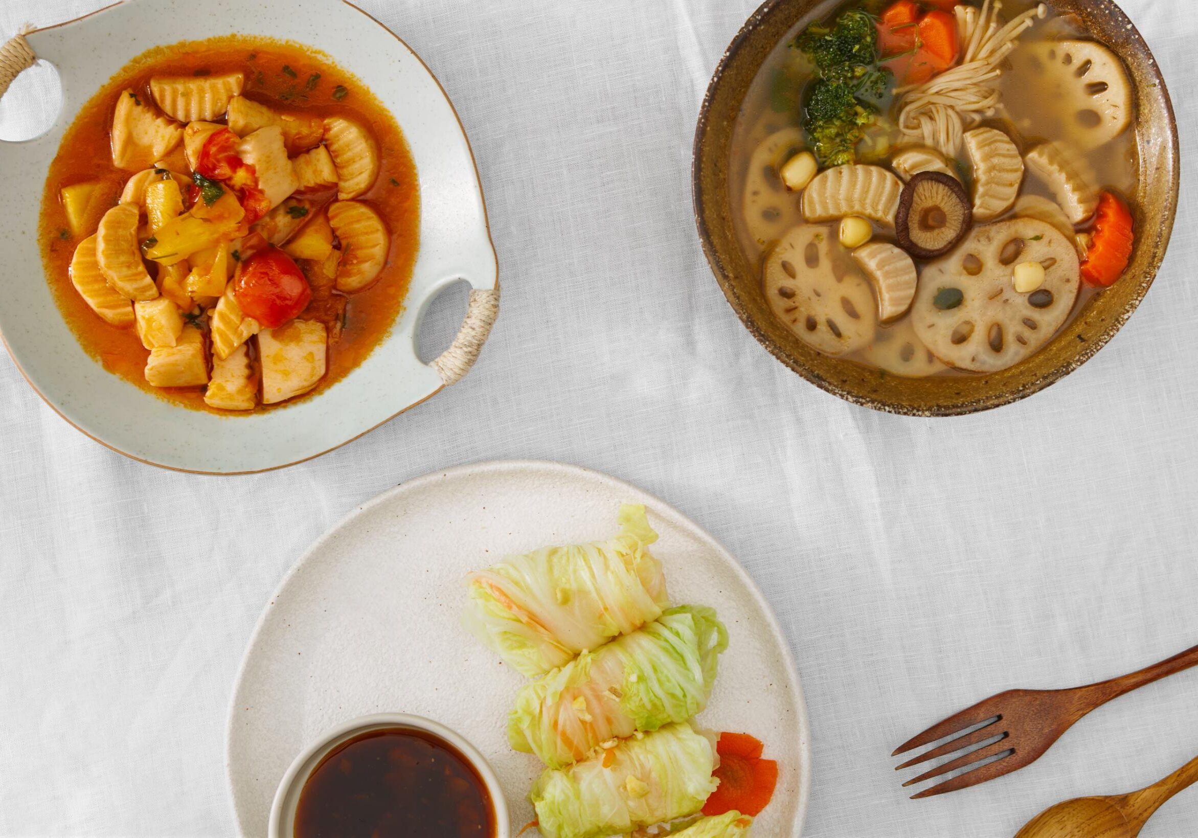 This image displays an assortment of vegetarian dishes arranged on a white tablecloth, featuring a main plate of purple rice accompanied by steamed broccoli, carrots, and tofu wrapped in seaweed. Surrounding the center are various side dishes, including a clear soup with lotus root and mushrooms, cabbage rolls, and a bowl of fresh greens.