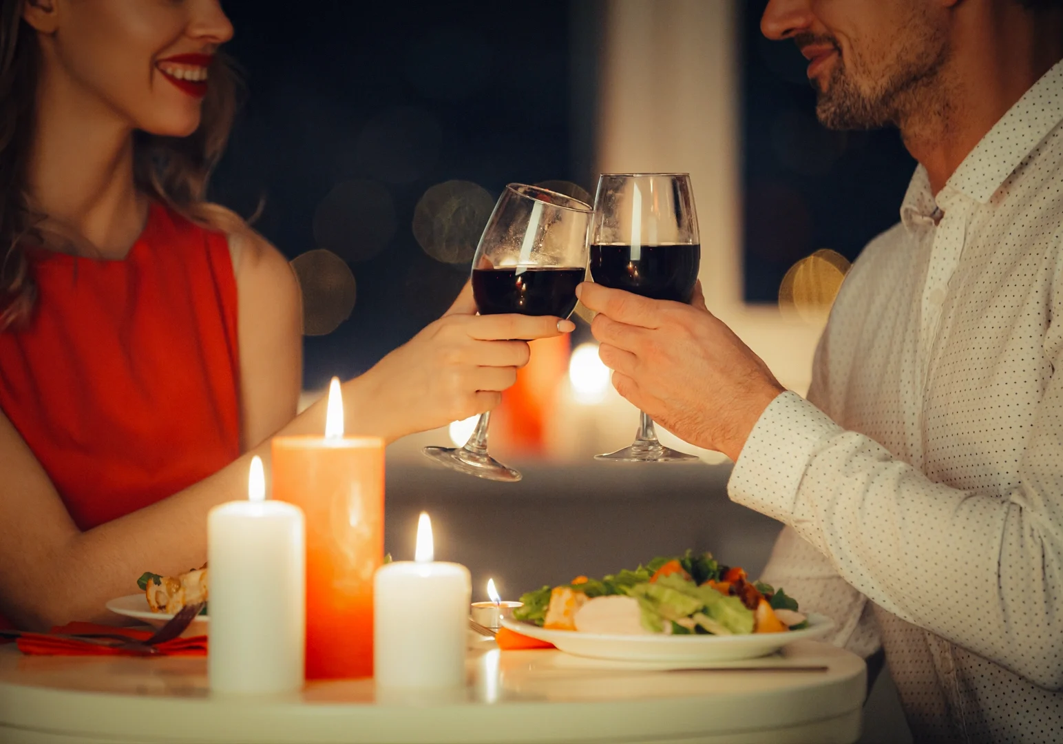 Close-up shot of a couple toasting red wine glasses at a candlelit dinner table with soft bokeh lights, romantic date night restaurant setting.