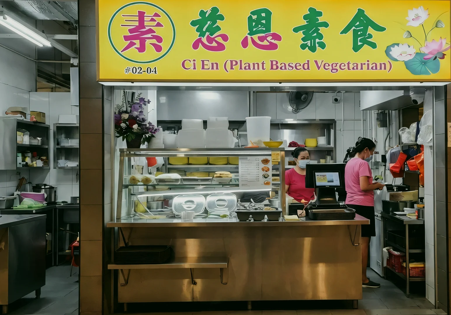 Eye‑level wide shot of a plant‑based vegetarian food stall with a yellow storefront sign reading “Ci En (Plant Based Vegetarian),” featuring a service counter, food display case, and kitchen preparation area behind.